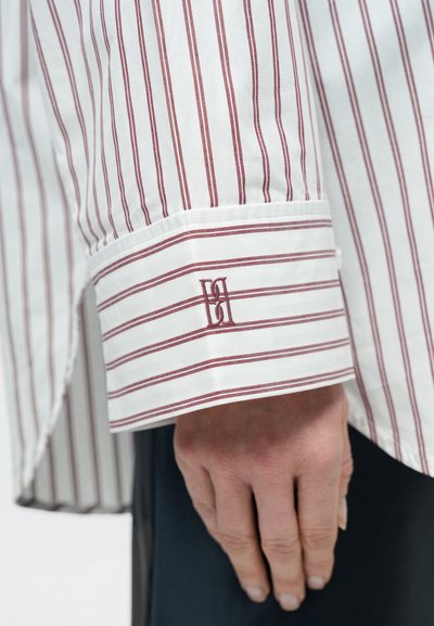Biela košeľa s červenými pásmi so zahnutým manžetou. Manžeta má červené vyšívané logo. Tkanina pôsobí hladko a sviežo.