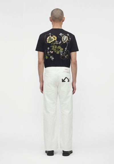 Άνδρας που φοράει μαύρο μπλουζάκι με πράσια και κίτρινη κεντητή floral διακόσμηση και λευκό παντελόνι με μαύρο βέλος στην πίσω τσέπη, στέκεται με την πλάτη γυρισμένη.