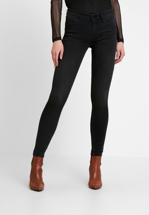 Femme portant un jean noir ajusté avec des poches avant et des bottines marron, debout avec une jambe légèrement pliée sur un fond blanc uni.
