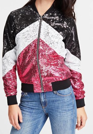 Femme portant une veste bomber brillante à sequins avec des blocs de couleurs noir, blanc et rouge, associée à un jean en denim bleu.