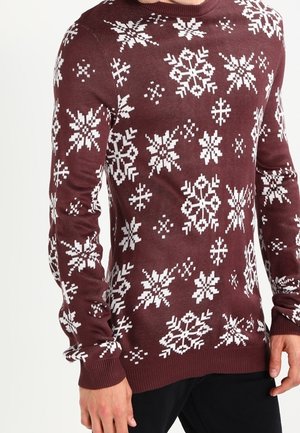 Vinrød sweater med hvide pixeliserede snefnugmønstre båret af en person med hænder afslappet langs siderne.
