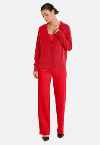 Rode cardigan met een V-hals, knoopsluiting en geribbelde manchetten. Gepaard met rode wijde broek en zwarte puntige schoenen.
