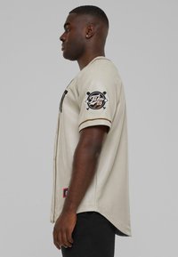 Maglia da baseball beige con texture in rete, accenti neri, logo "Tabus 05" sulla manica sinistra e orlo curvo.