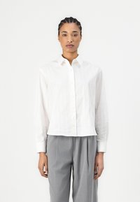 Camicia bianca con righe verticali, maniche lunghe e colletto classico, abbinata a pantaloni grigi con pieghe. Texture liscia.