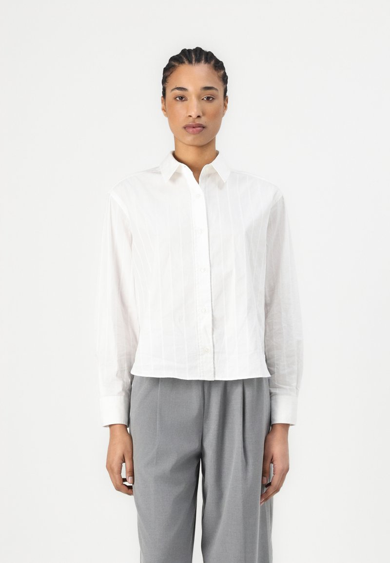 Camicia bianca con righe verticali, maniche lunghe e colletto classico, abbinata a pantaloni grigi con pieghe. Texture liscia.