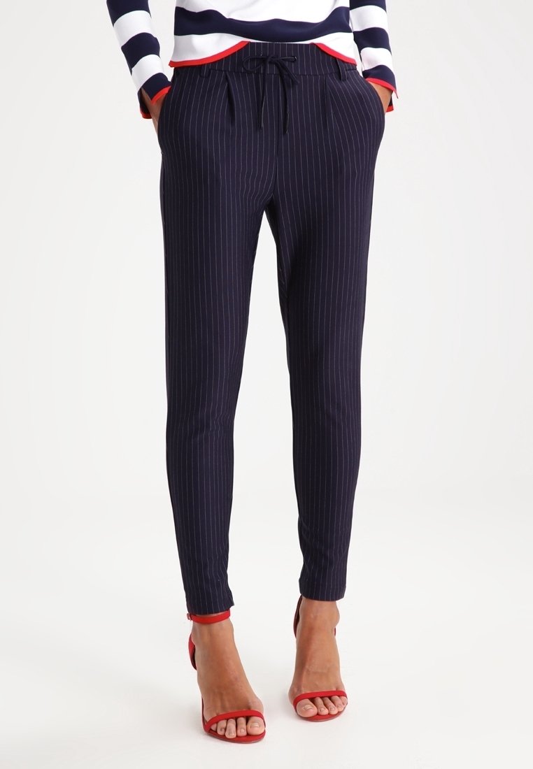 Femme portant un pantalon fuselé bleu marine à fines rayures avec une taille à cordon et des sandales rouges à talons hauts, mains dans les poches.