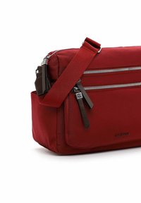 Červená nylonová crossbody taška s dvěma předními zipovými kapsami, hnědými koženými akcenty a nastavitelním popruhem. Má obdélníkový tvar a hladkou texturu.