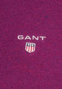 Lila Strickpullover-Stoff mit dem Schriftzug "GANT" und einem Logo, das aus einem Schild mit roten und weißen Streifen sowie Sternen besteht.