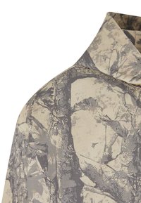 Vaaleanbeige kauluspaita, jossa on tummanharmaa puunrungon kuvio. Kangas on hienovaraisesti teksturoitu, mikä korostaa luonnollista designia.