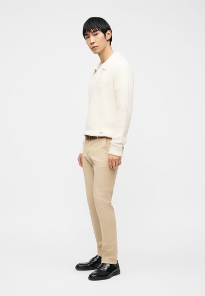 Άνδρας όρθιος, φοράει λευκό πλεκτό πουλόβερ με μακριά μανίκια, μπεζ παντελόνι με καφέ ζώνη και μαύρα δερμάτινα παπούτσια, με φόντο απλό λευκό.