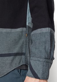 Gros plan sur le bras d'une personne portant une chemise à manches longues rayée noir et gris avec une petite étiquette orange BOSS sur la couture latérale.