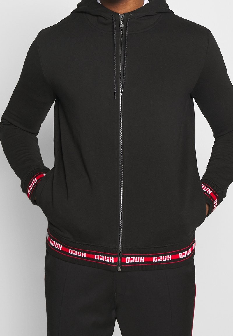 Homme portant un sweat à capuche noir zippé avec des bandes de logo rouges et blanches aux poignets et à l'ourlet, mains dans les poches, debout devant un fond clair.