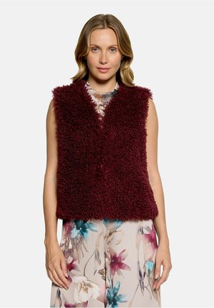 Frau trägt eine ärmellose bordeauxrote flauschige Weste über einem Kleid mit floralem Muster in Blau-, Pink- und Beigetönen, stehend vor weißem Hintergrund.