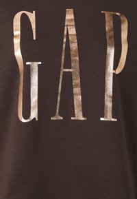 Camiseta de algodón marrón con grandes letras metálicas doradas que dicen "GAP" en la parte frontal, con una textura suave y un corte clásico.
