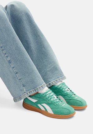 Zielone zamszowe tenisówki z białymi akcentami i gumową, brązową podeszwą; posiadają sznurowadła oraz niski profil. Noszone z jasnoniebieskimi spodniami dżinsowymi.