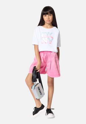 Meisje met lang donker haar, draagt een wit DKNY T-shirt, roze korte broek, zwart-witte sneakers en houdt een zilveren DKNY crossbodytas vast.