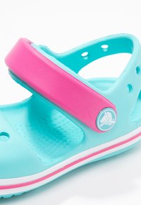 Turkoosi avonainen sandaali, jossa on kirkkaan vaaleanpunainen hihna. Varustettu pienillä ilmastointia varten tehdyillä rei’illä ja sivulla pyöreällä logolla. Pehmeä, teksturoitu materiaali.