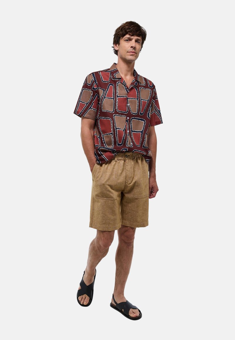 Chemise à manches courtes avec un motif abstrait rouge et beige, associée à un short beige. Portée avec des sandales noires. Tissu léger et coupe décontractée.