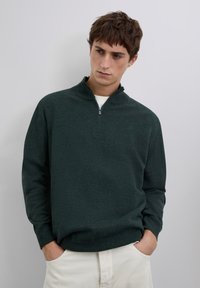 Maglione verde scuro a collo alto in tessuto morbido lavorato a maglia, con collo con zip e maniche lunghe. Design minimalista senza motivi visibili.