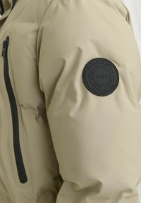 Beige puffer jacket with a textured exterior, featuring a black zip pocket and circular black logo patch on the left shoulder.