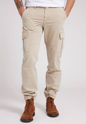 Hombre con pantalones cargo beige con bolsillos laterales y botas de ante marrones con cordones, con las manos en los bolsillos, de pie sobre un fondo liso.