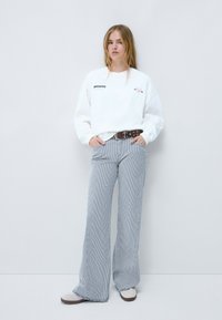 Sudadera blanca con texto negro y detalles gráficos, combinada con pantalones de pierna ancha a rayas. Cinturón marrón con herrajes plateados y zapatos beige.