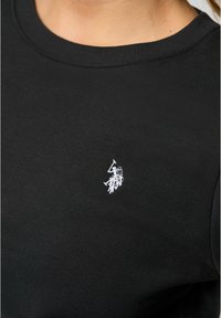 Svart bomullströja med rund halsringning och ribbade ärmslut, med en liten vit broderad logotyp på vänster bröst.