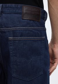 Κοντινό της μέσης ενός ατόμου που φορά σκούρα μπλε τζιν με ορατή πίσω τσέπη και καφέ δερμάτινη ετικέτα Diesel.