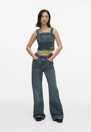 Giovane donna in piedi con le mani in tasca, indossa un top verde a strati e jeans larghi in denim scuro con un top corto in denim abbinato.