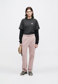 Νέος ενήλικας που φοράει μαύρο πουκάμισο με μακριά μανίκια και μπλούζα, ροζ φόρμες, γκρι αθλητικά παπούτσια, κρατώντας ένα μπεζ καπέλο μπροστά από λευκό φόντο.