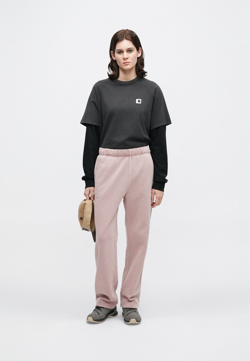 Giovane adulto che indossa una maglietta a maniche lunghe nera a strati e una t-shirt, pantaloni della tuta rosa, scarpe grigie, tenendo un berretto beige su uno sfondo bianco.