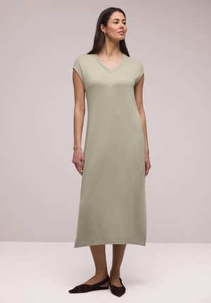 Frau steht und trägt ein ärmelloses, hellolivfarbenes Midikleid mit V-Ausschnitt und braunen flachen Slingback-Schuhen vor neutralem Hintergrund.