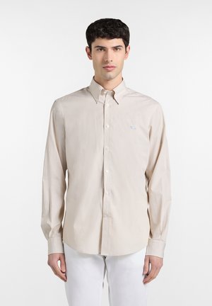 Junger Mann trägt ein beiges langärmeliges Hemd mit Knöpfen und weiße Hose, steht vor einem einfachen weißen Hintergrund.