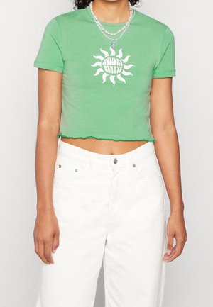 Zielony, krótki t-shirt z bawełny, z białym wzorem słońca. Surowy brzeg u dołu oraz krótkie rękawy w połączeniu z białymi spodniami o wysokim stan.