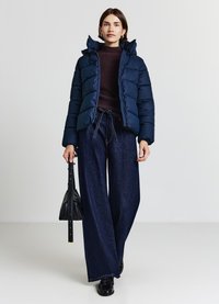 Giacca imbottita blu navy con cappuccio, caratterizzata da un'imbottitura trapuntata. Indossata sopra un maglione scuro e abbinata a pantaloni in denim scuro a gamba larga. Borsa nera.