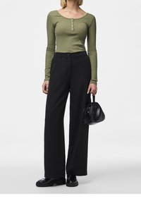 Kvinna i en olivgrön långärmad ribbad topp, svarta vida byxor, svarta skor, håller en liten svart handväska mot en vit bakgrund.