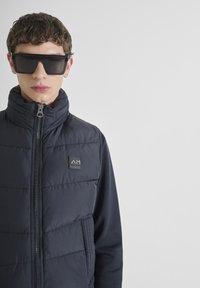 Chaleco de plumas navy con cuello alto, cremallera frontal y parche con logo. El modelo lleva gafas de sol negras angulares. Telas suaves y texturizadas.