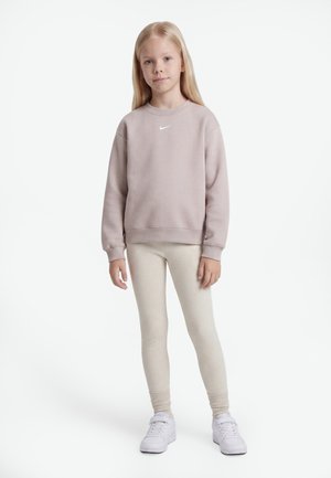 Mladé dievča s dlhými blond vlasmi, ktoré má na sebe svetloružovú mikinu Nike, krémové rebrované legíny a biele tenisky, stojace na bielom pozadí.