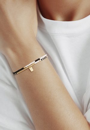Arm, der zwei dünne Perlenarmbänder in Schwarz, Pink und Gold mit einem kleinen rechteckigen goldenen Anhänger trägt, vor einem weißen Hemdhintergrund.