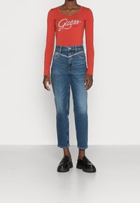 Rode longsleeve bodysuit met "Guess" logo; hoog-onafhankelijk blauwe jeans met rechte snit; zwarte instappers met grove zolen.