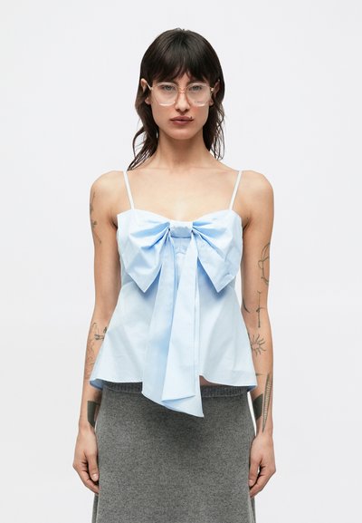 Haut en coton bleu clair avec un grand nœud à l'avant, bretelles fines et silhouette évasée, associé à une jupe grise. Texture lisse.