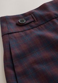 Gros plan d’un tissu à carreaux bleu et bordeaux avec un détail de languette boutonnée sur la ceinture d’un vêtement sur mesure.