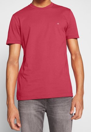 Man draagt een effen rood T-shirt met korte mouwen en ronde hals met een klein wit "ck"-logo op de linkerborst en grijze jeans.