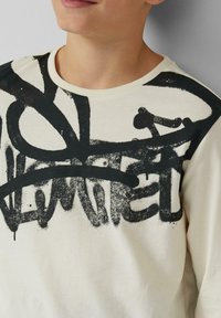 Kremowy bawełniany sweatshirt z dużym czarnym nadrukiem w stylu graffiti na klatce piersiowej i ramieniu, z żebrowanym dekoltem i długimi rękawami.