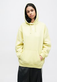 Svetložltá mikina s kapucňou vyrobená z mäkkej látky, s predným vreckom a sťahovacou šnúrkou na kapucni. Minimalistické biele logo nad vreckom. Pohodlný strih.