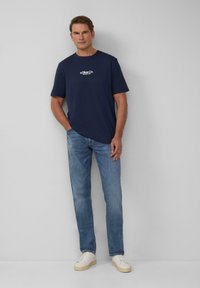 Marineblauw T-shirt met ronde hals en wit logo, gecombineerd met lichtblauwe rechte jeans en witte sneakers met beige zolen.