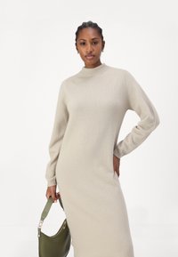 Robe en tricot beige avec un col montant, des manches longues et des bords côtelés, accompagnée d'un sac à main vert avec une bandoulière.