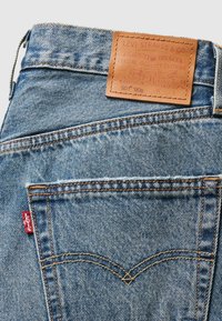 Gros plan sur la poche arrière d'un jean en denim Levi's bleu clair avec des surpiqûres orange, une étiquette rouge Levi's et un patch de marque en cuir beige sur la ceinture.