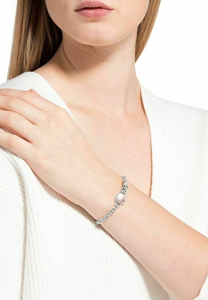Femme portant un haut blanc texturé avec un bracelet chaîne argenté orné d’une pierre rose ovale unique sur son poignet.