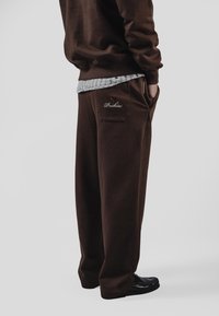 Braune Jogginghose aus weichem Stoff, mit einer Gesäßtasche, die das rote bestickte "Pockies"-Logo trägt, sowie seitlichen Taschen.